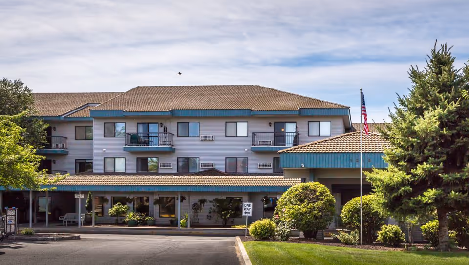Exterior view of a three-story senior living facility building with balconies, a covered entrance, well-maintained landscaping including bushes and trees, and an American flag on a flagpole in front.