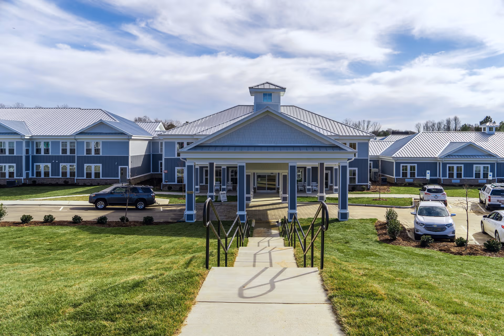 Front exterior view of Arbor Ridge at Huntersville, showing a large building with a covered entrance, metal roof, multiple windows, a concrete walkway with handrails leading up to the entrance, and several parked cars on either side. The sky is partly cloudy.