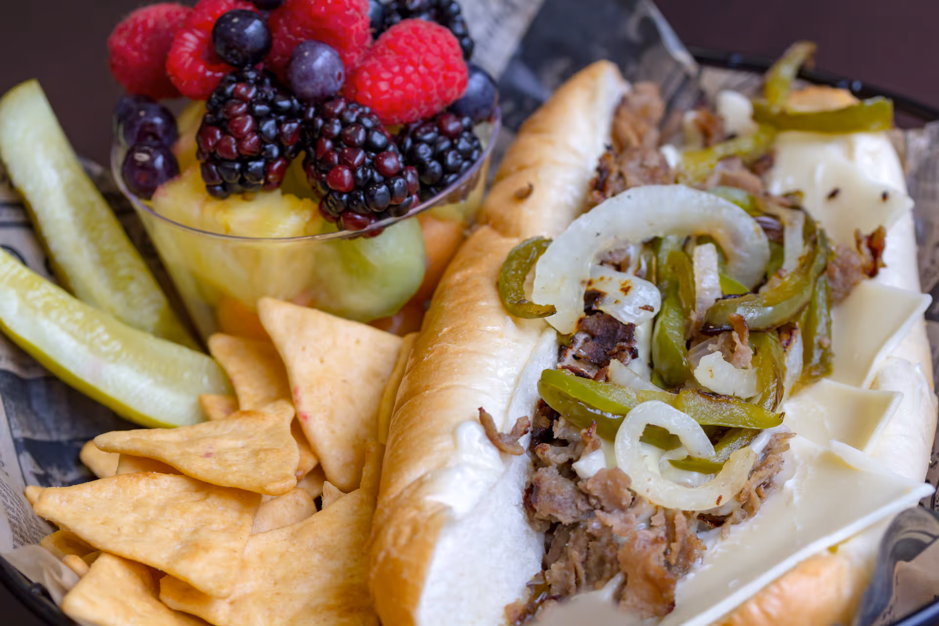 Close-up of a sandwich with melted cheese, grilled onions and peppers served with crackers, pickle spears and a cup of mixed berries.