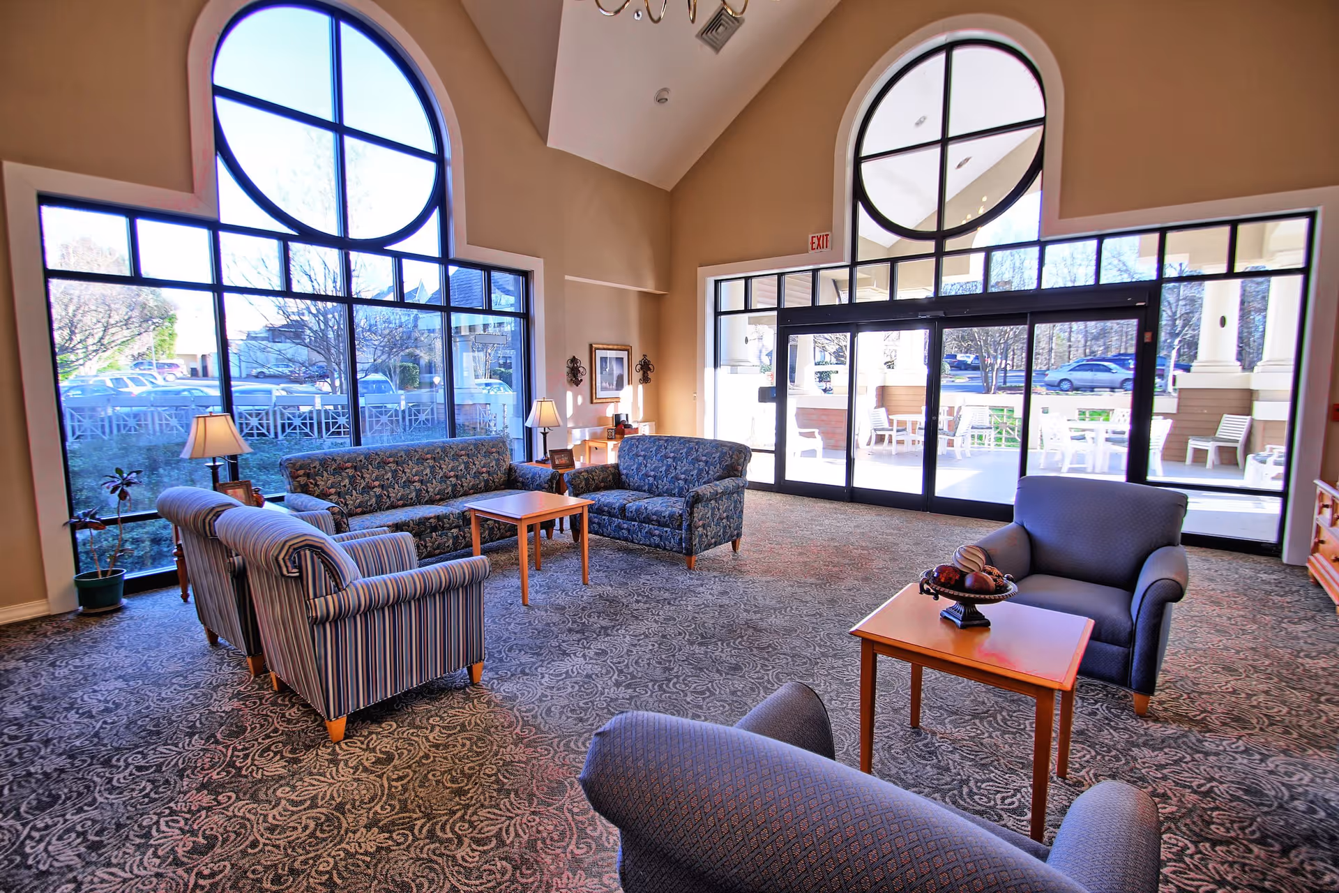 A spacious and well-lit living room area with large arched windows and glass doors letting in natural light. The room is furnished with patterned sofas, armchairs, and wooden coffee tables. There are table lamps and decorative items on the tables, and outside the glass doors, there is a patio area with white chairs and tables.