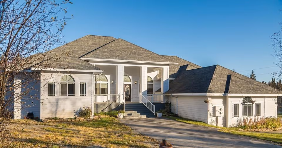 Front exterior view of a single-story assisted living home with white siding, multiple windows with arched tops, a central entrance with steps and a small porch, surrounded by a grassy yard and clear blue sky.