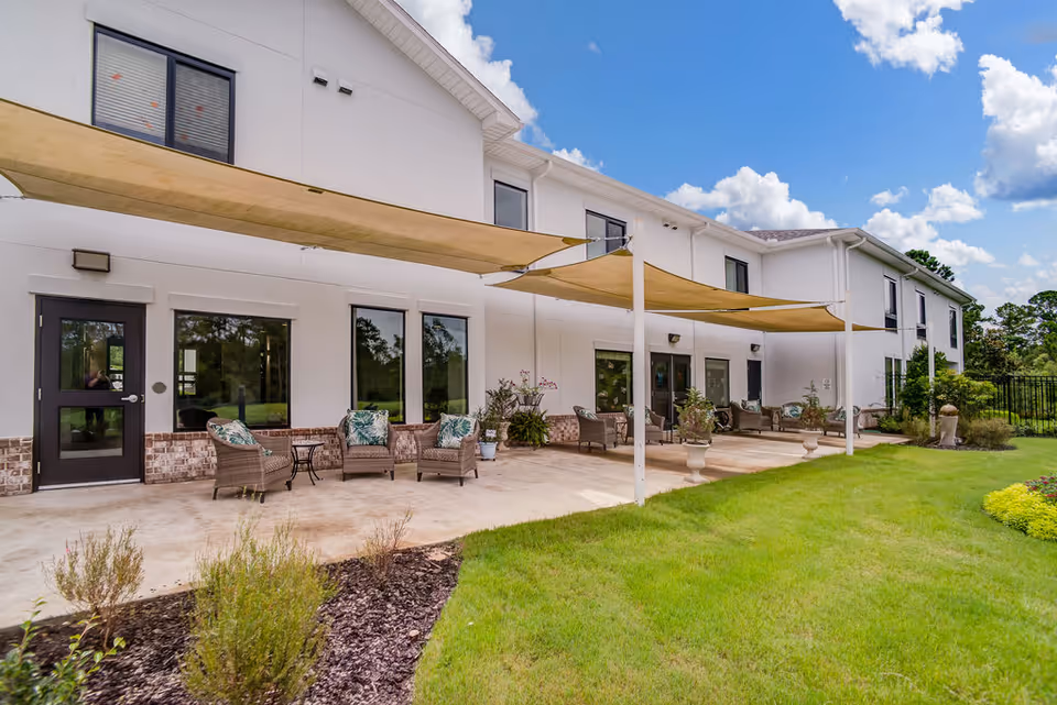 Outdoor patio area of a senior living facility with wicker chairs and small tables arranged under beige shade sails attached to the building. The building is white with large windows and a well-maintained lawn and garden beds in the foreground under a partly cloudy blue sky.