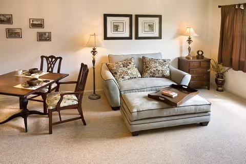 A cozy living room and dining area with a beige loveseat and matching ottoman, decorated with patterned cushions and a wooden tray on the ottoman. There are two framed pictures on the wall above the loveseat, a wooden side table with a lamp and decorative items, and a floor lamp next to the loveseat. The dining area has a wooden table set with placemats, bowls, and cups, accompanied by two wooden chairs with cushioned seats. The room has beige carpeting and a window with brown curtains.