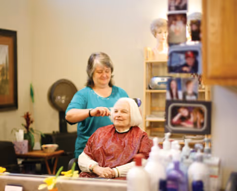 A senior woman with white hair sits in a salon chair wearing a red cape while a hairstylist stands behind her, combing her hair. The setting appears to be a hair salon with various hair care products and a mirror reflecting the scene.