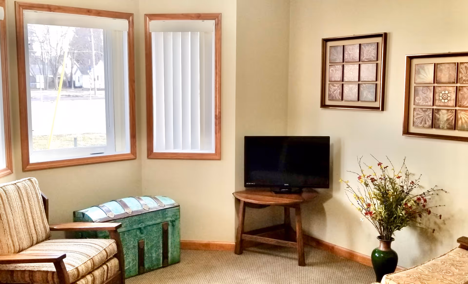 A cozy corner of a living room with two windows featuring vertical blinds, a striped armchair, a green decorative chest, a small wooden table with a flat-screen TV, a vase with colorful flowers, and two framed wall art pieces.