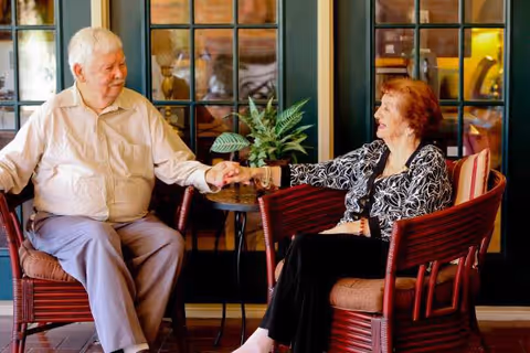 An elderly man and woman sitting in cushioned wooden chairs facing each other, holding hands and smiling. They are indoors in front of glass-paneled doors with a small table and a potted plant between them.