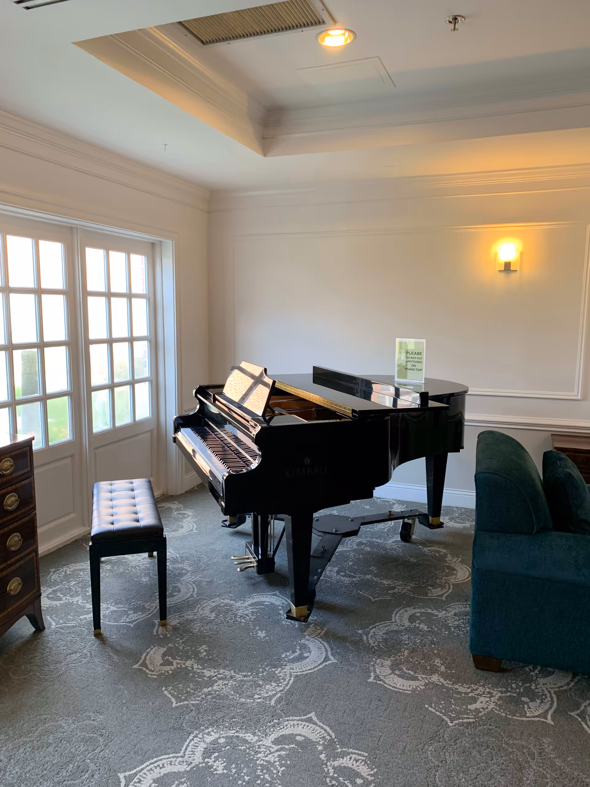 A bright room with a black grand piano and a matching padded bench on a patterned carpet. The room has white walls with molding, a large window with multiple panes letting in natural light, a green upholstered sofa, a wooden chest of drawers, and a wall-mounted light fixture.
