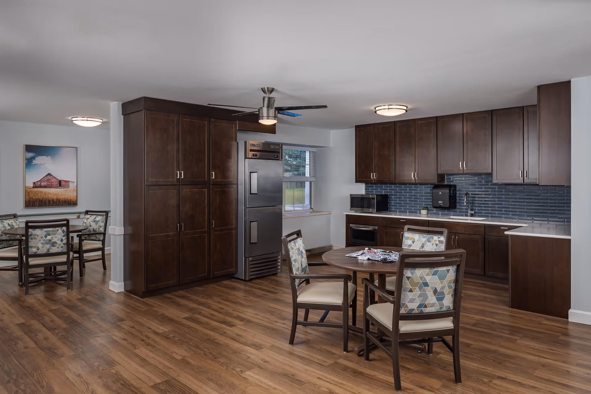 A communal kitchen and dining area with dark wood cabinets, a round table and chairs, and hardwood floors.