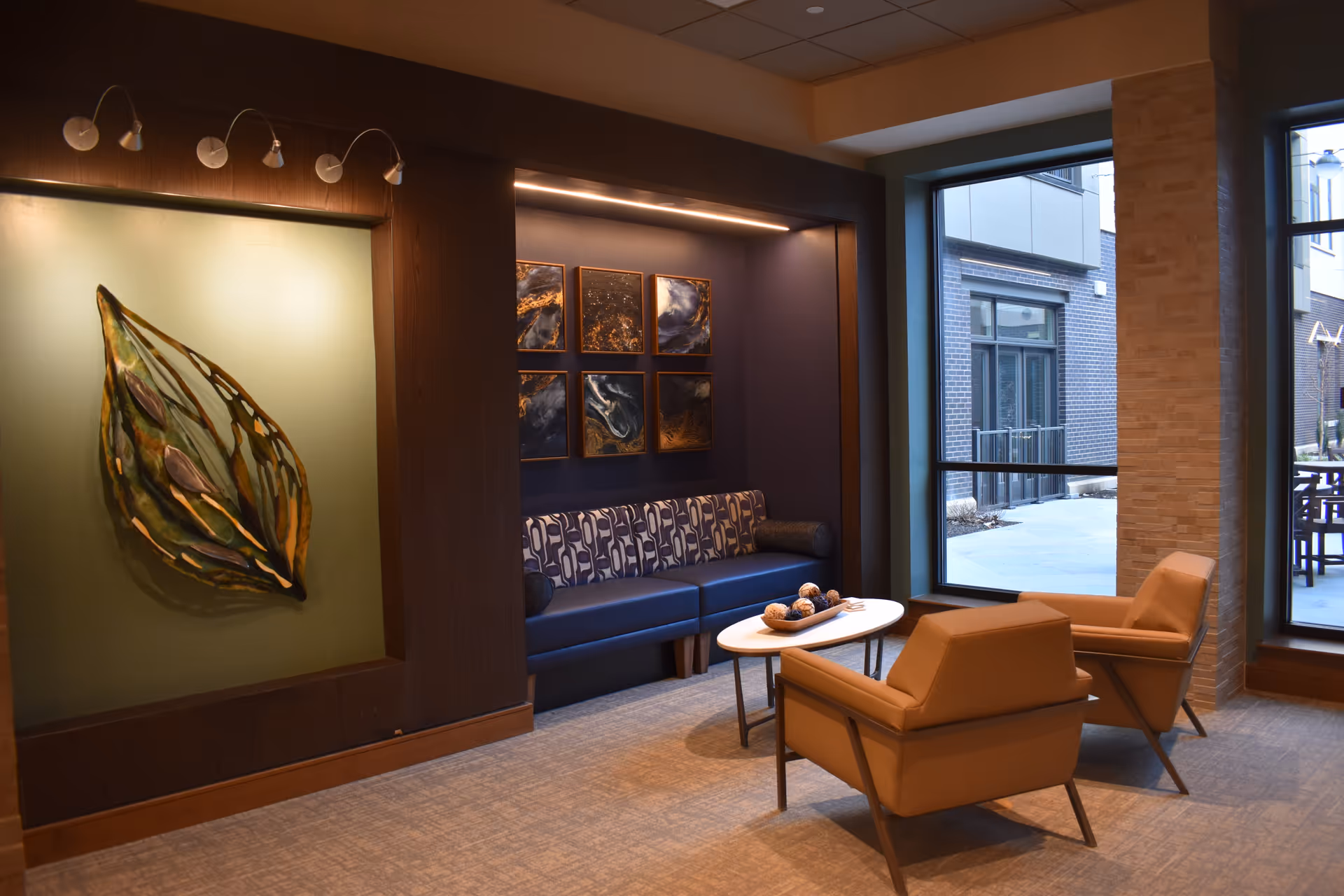 A modern lounge area with a blue patterned sofa, two tan armchairs, and a white oval coffee table with decorative items. The wall behind the sofa features six abstract art pieces, and a large sculptural leaf artwork is displayed on the adjacent wall. Large windows provide a view of an outdoor patio area.