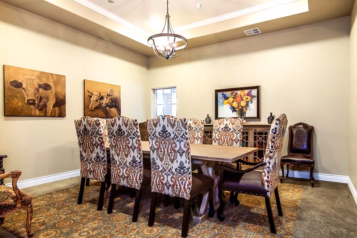 A dining room with a wooden table surrounded by eight upholstered chairs with patterned fabric. The room has beige walls adorned with three paintings: two featuring cows and one with a colorful bouquet of flowers. A chandelier hangs from the ceiling, and a wooden sideboard with decorative items is against the far wall.