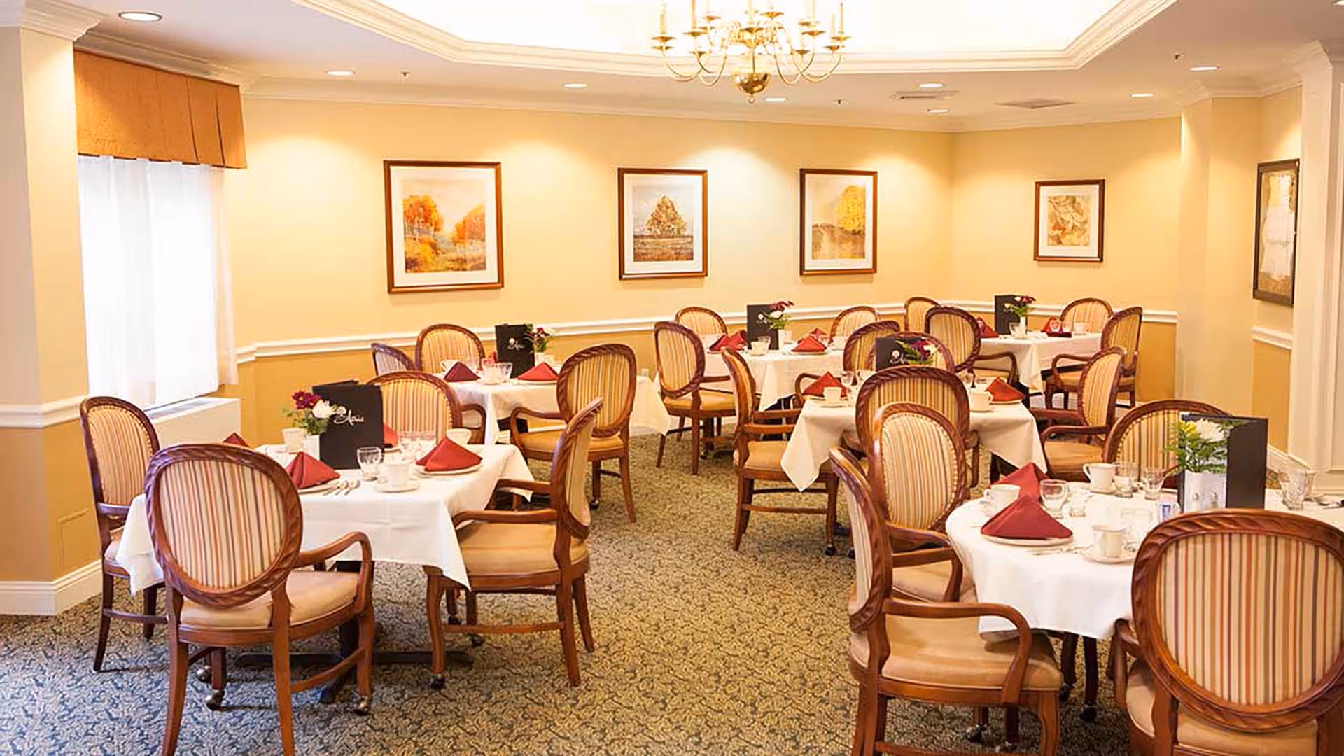 A well-lit dining room with multiple round and square tables covered with white tablecloths, each set with red folded napkins, cups, glasses, and small flower arrangements. The room has beige walls adorned with framed artwork and a chandelier hanging from the ceiling. The chairs have wooden frames with striped upholstery.