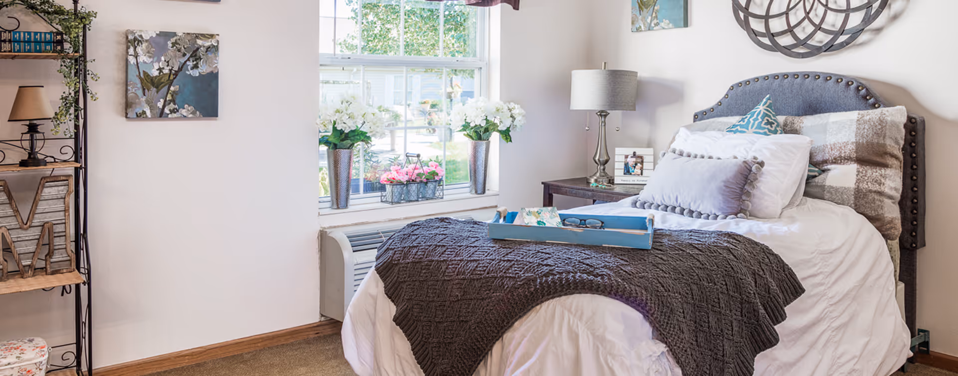 A cozy bedroom with a neatly made bed featuring multiple pillows and a dark knitted blanket. A blue tray with glasses and a book rests on the bed. Next to the bed is a wooden nightstand with a lamp and a framed photo. A large window behind the bed lets in natural light and has white flowers on the windowsill. On the left side, there is a metal and wood shelving unit with decorative items and books. The walls are light-colored with floral artwork and a decorative round mirror above the bed.