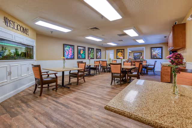 A spacious dining room with several tables and chairs arranged neatly. The room features wood flooring, beige walls, and ceiling lights. There are colorful framed artworks on the far wall and a large aquarium built into a white cabinet on the left side. A 'WELCOME' sign is displayed above the aquarium. Fresh flowers in vases are placed on the tables and countertop.