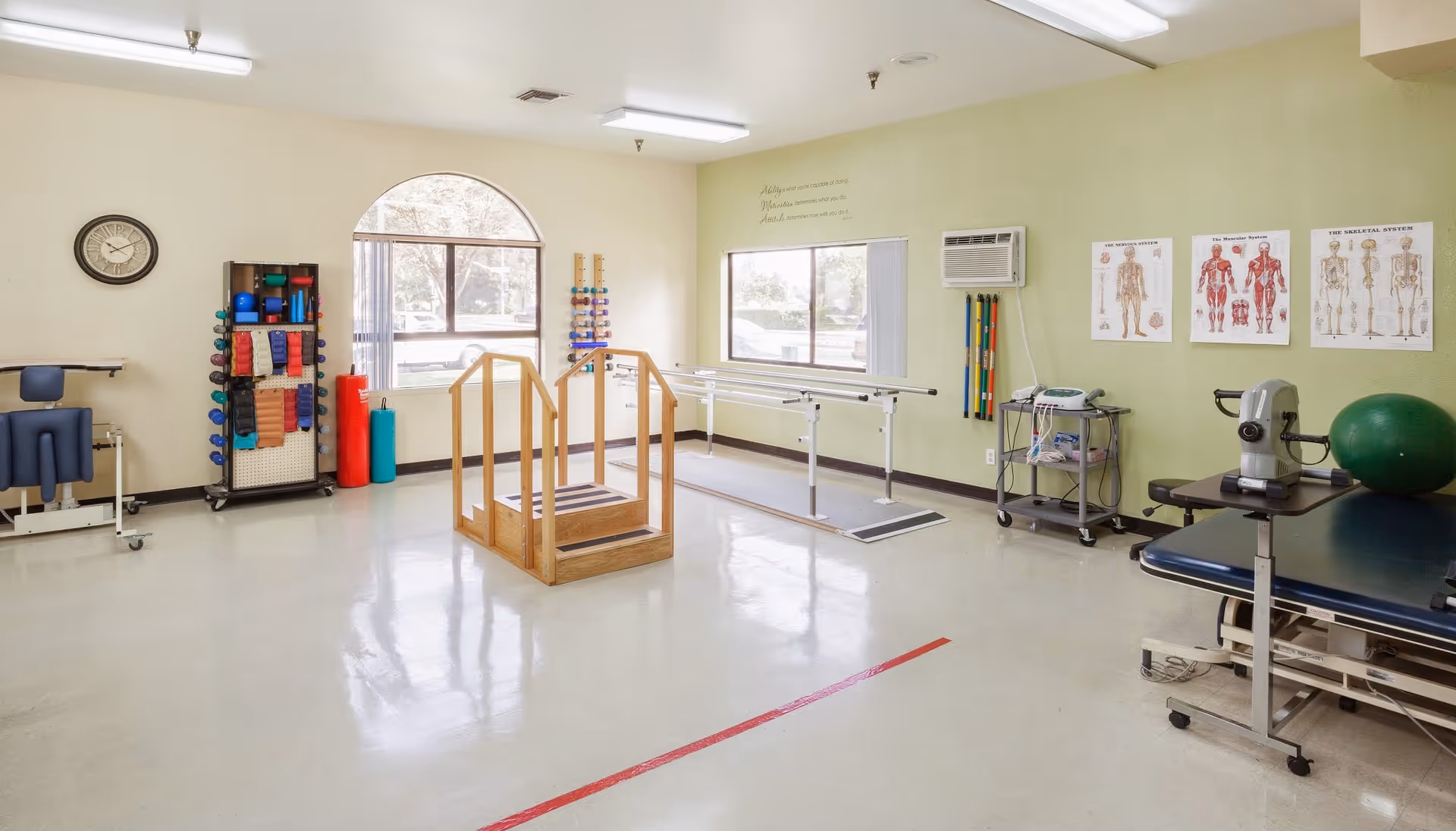 A physical therapy room with exercise equipment including parallel bars, a wooden step platform, various weights and exercise balls, anatomical charts on the wall, and a treatment table. The room has large windows letting in natural light and a clock on the wall.