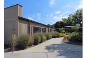 Exterior view of a single-story building with brown siding, large windows, and a concrete walkway surrounded by bushes and greenery under a blue sky with some clouds.