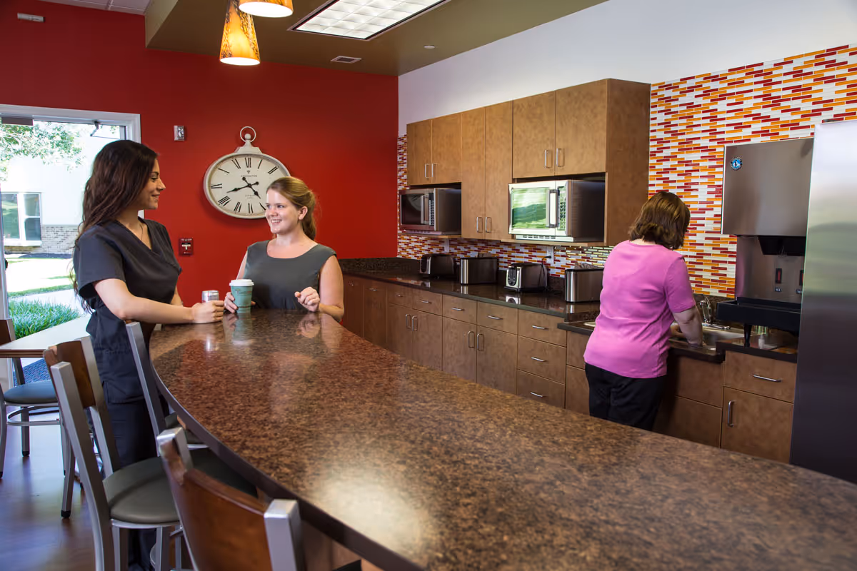 Three women talking and preparing beverages at a long counter in a communal kitchen with wood cabinets and a colorful tile backsplash.