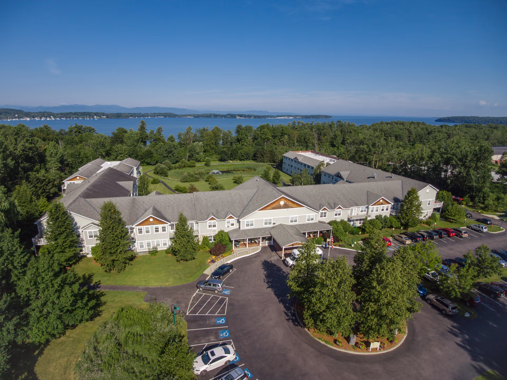 Aerial view of The Residence at Shelburne Bay senior living facility surrounded by trees and greenery, with a large parking lot in front and a lake visible in the background under a clear blue sky.