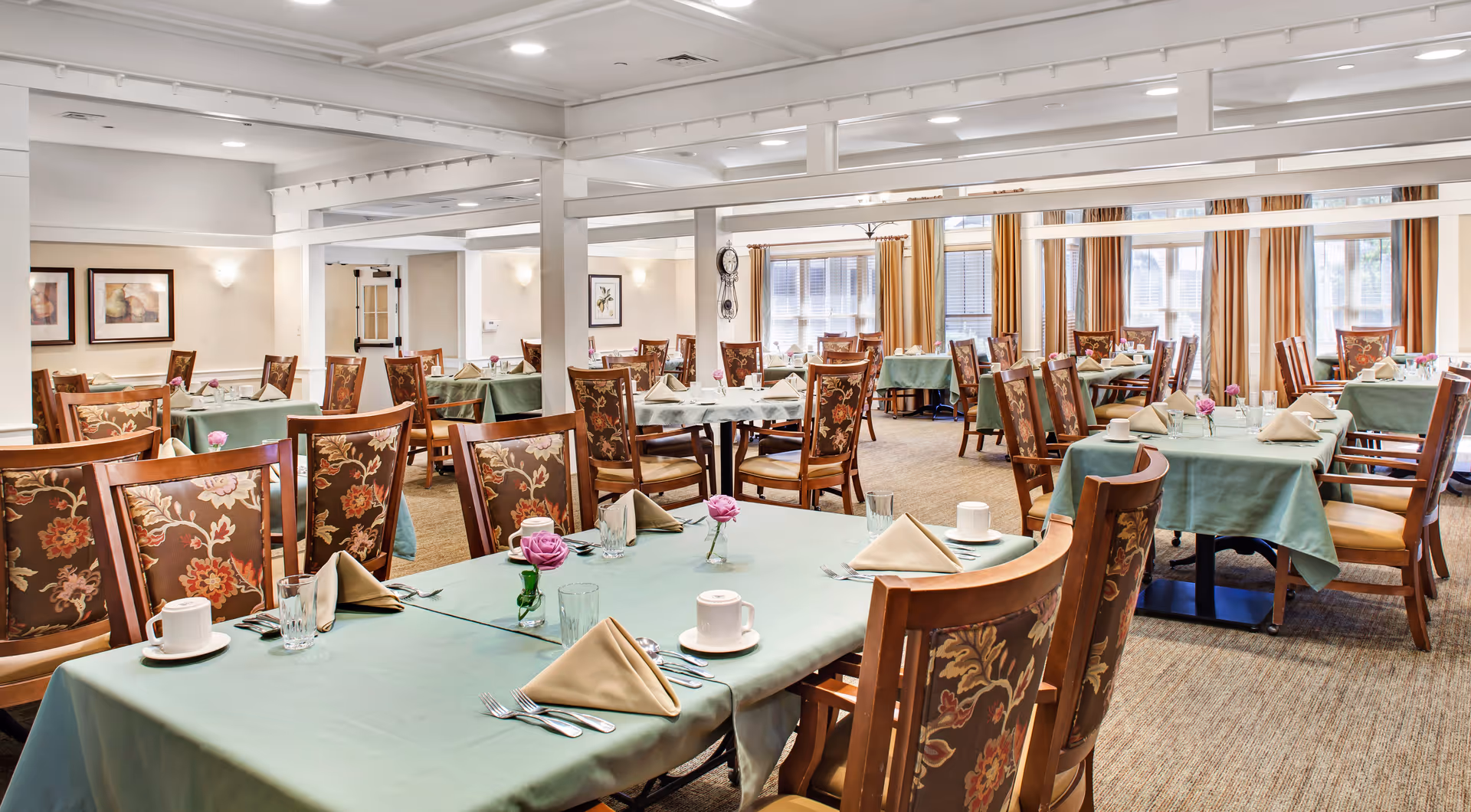 Spacious dining room with multiple tables set with green tablecloths, folded napkins, floral centerpieces and wooden chairs.