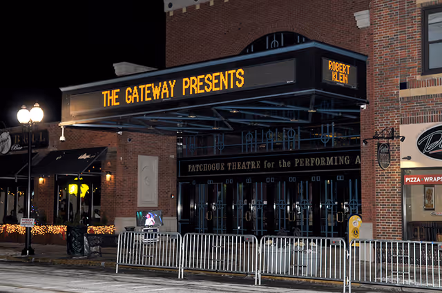 Nighttime exterior view of the Patchogue Theatre for the Performing Arts with a marquee displaying 'THE GATEWAY PRESENTS' and 'ROBERT KLEIN'. The building is made of brick and there are metal barricades in front of the entrance. Nearby street lamps and adjacent businesses are also visible.