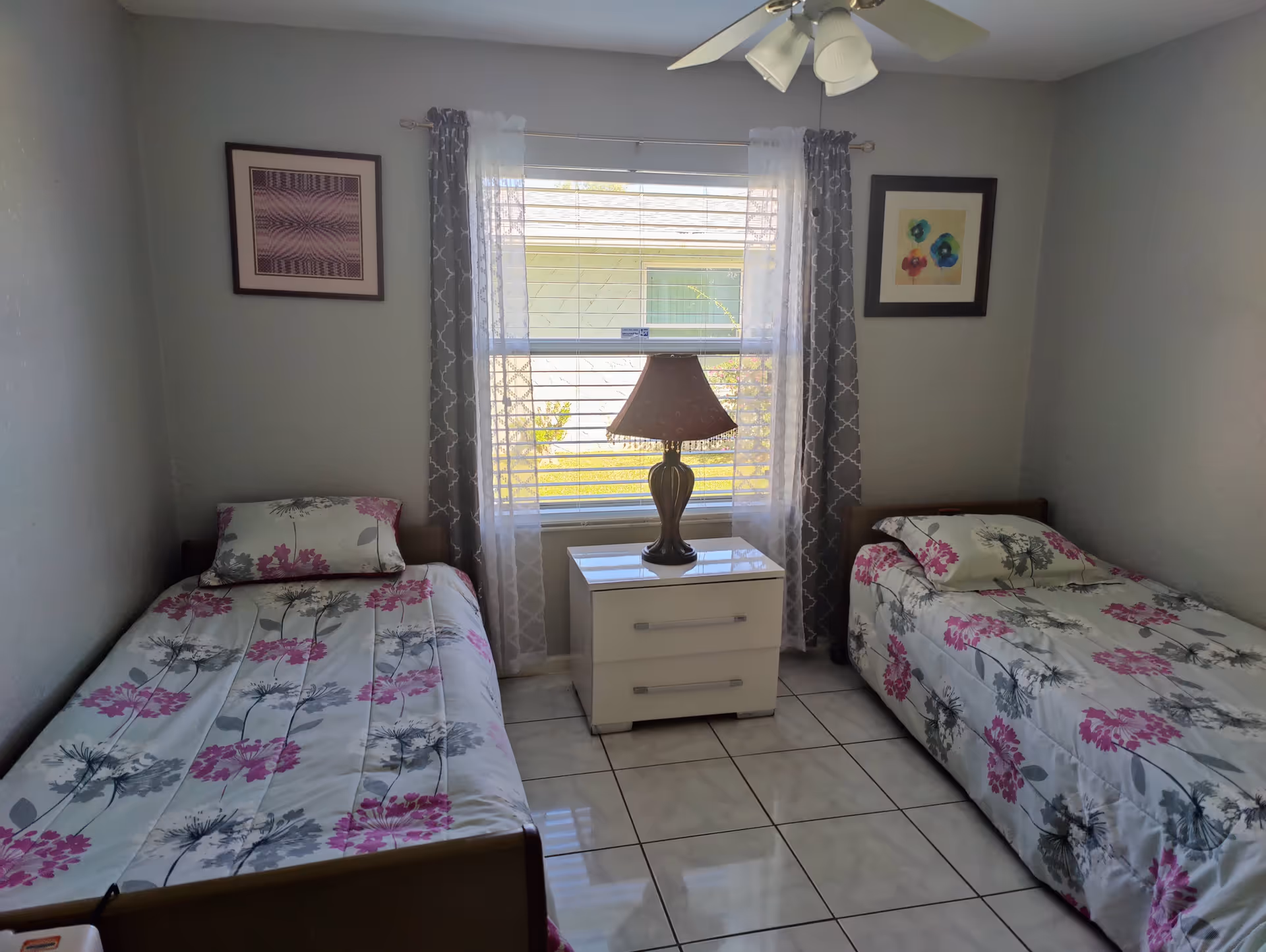 A small bedroom with two single beds on either side of a white nightstand with a lamp. The beds have matching floral bedspreads in pink and gray. A window with sheer white and patterned gray curtains is centered on the wall behind the nightstand. Two framed pictures hang on the walls above each bed. The floor is tiled and a ceiling fan with lights is visible above.