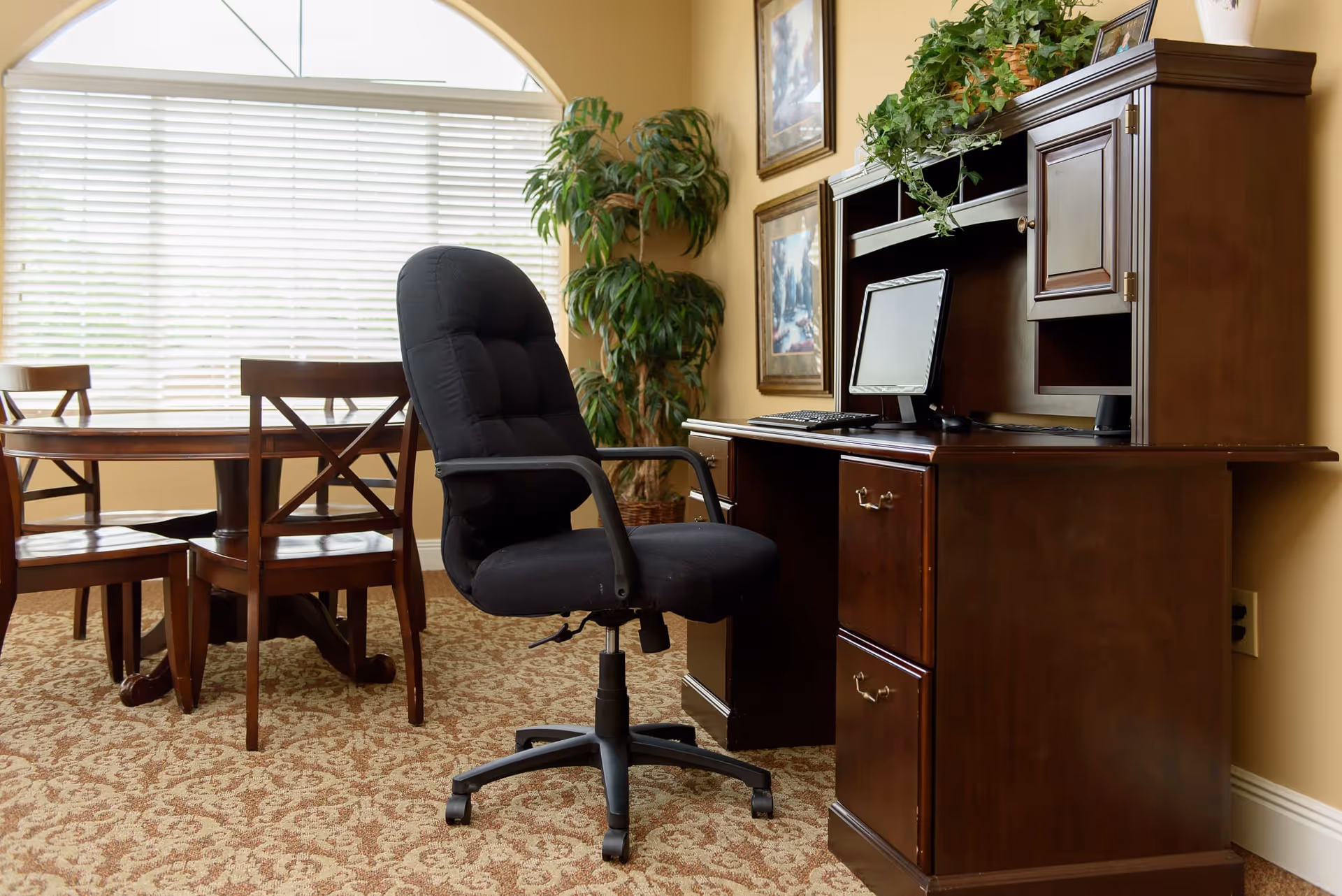A cozy office area with a dark wooden desk and hutch, a black cushioned office chair on wheels, a computer monitor and keyboard on the desk, a potted plant on top of the hutch, and a dining table with four wooden chairs near a large window with white blinds.