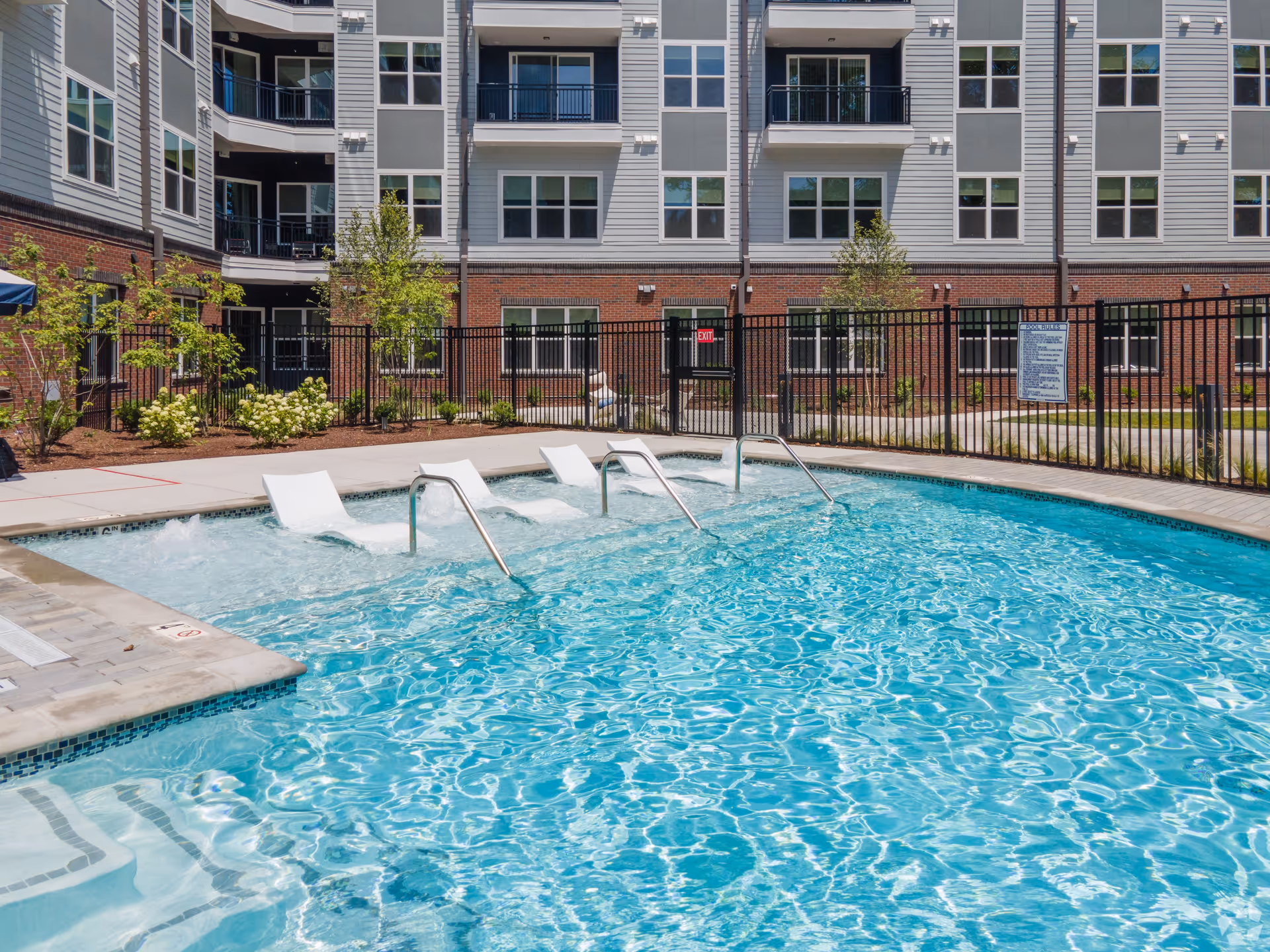 Outdoor swimming pool with built-in lounge chairs and metal handrails, surrounded by a black metal fence. Behind the pool is a multi-story residential building with balconies and windows, and landscaped greenery along the fence.