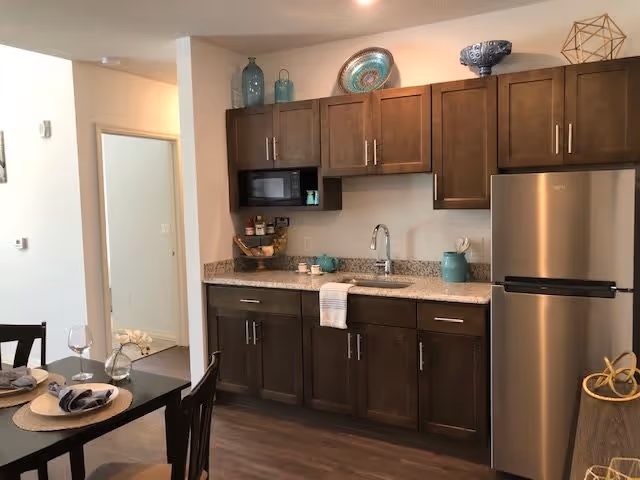 Open kitchenette with dark wood cabinets, granite countertop, stainless steel refrigerator, sink, and a nearby dining table set for two.
