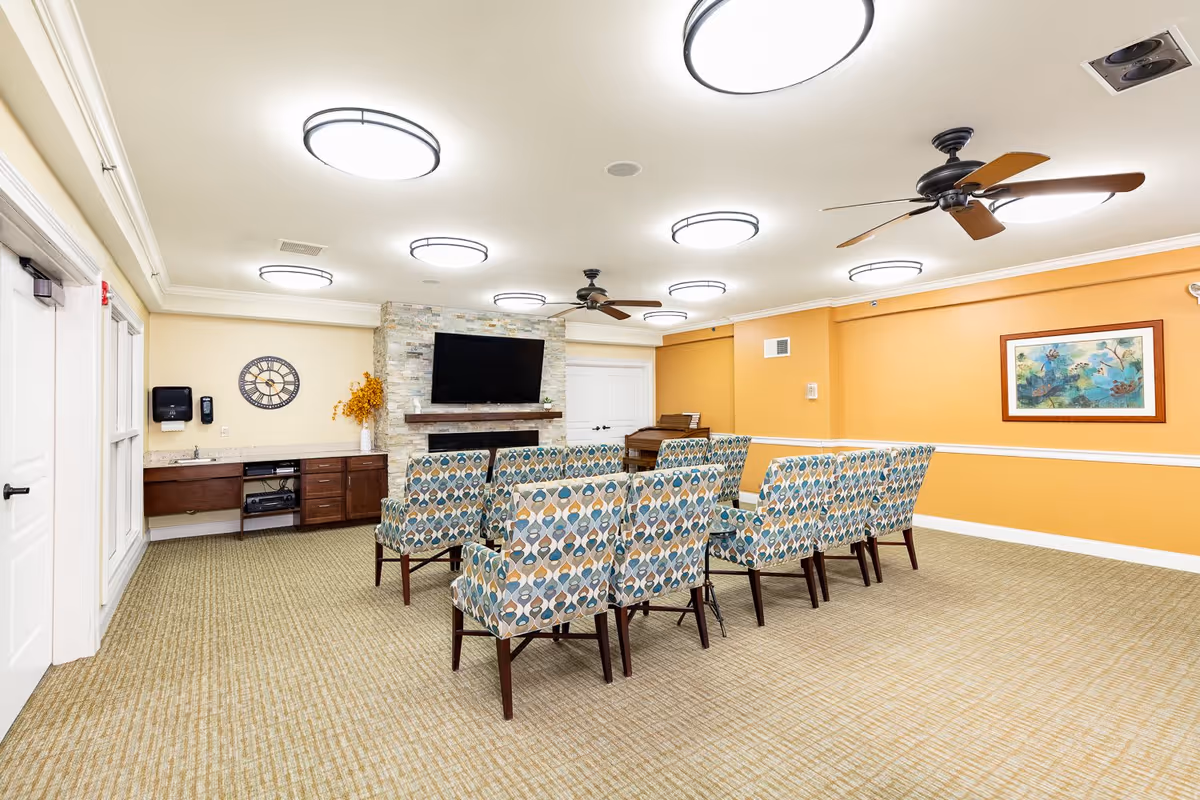 A well-lit common room with patterned upholstered chairs arranged in rows facing a wall-mounted flat-screen TV above a stone fireplace. The room has beige carpet, yellow and white walls, ceiling fans, and round ceiling lights. There is a wooden cabinet with a sink and a clock on the left wall, and a framed painting on the right wall.