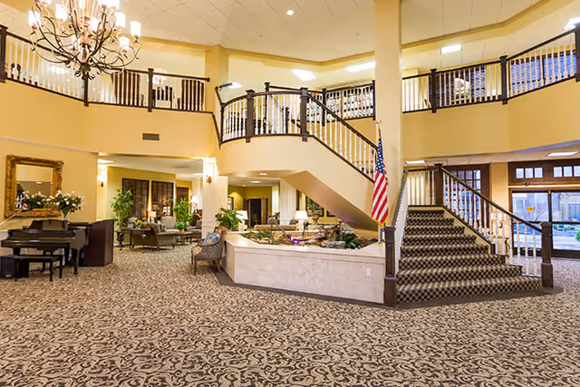 Spacious senior living facility lobby with a large patterned carpet, a grand staircase with dark wooden railings, an American flag, a chandelier, a piano, and seating areas with plants and lamps. The upper level has a balcony overlooking the lobby.