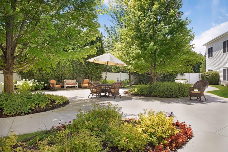 Outdoor patio area with a round table and umbrella surrounded by four wicker chairs, additional seating with cushioned chairs and a bench, lush green trees and shrubs, and a white building in the background under a partly cloudy sky.