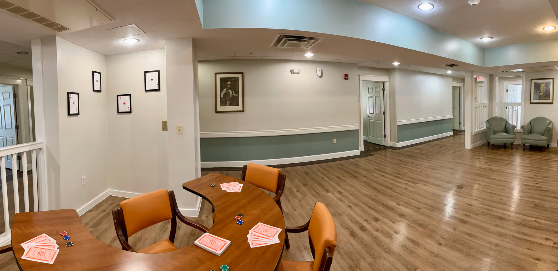A spacious interior room with wood flooring and light-colored walls. There is a curved wooden table with orange cushioned chairs around it, playing cards and poker chips are placed on the table. The walls have framed pictures, including large playing cards and a portrait. Two green armchairs are positioned near the far wall, and several doorways lead to other rooms.