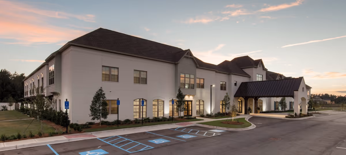 Exterior view of a two-story senior living facility building at dusk with a covered entrance, several windows, landscaped greenery, and multiple handicap parking spaces in front.