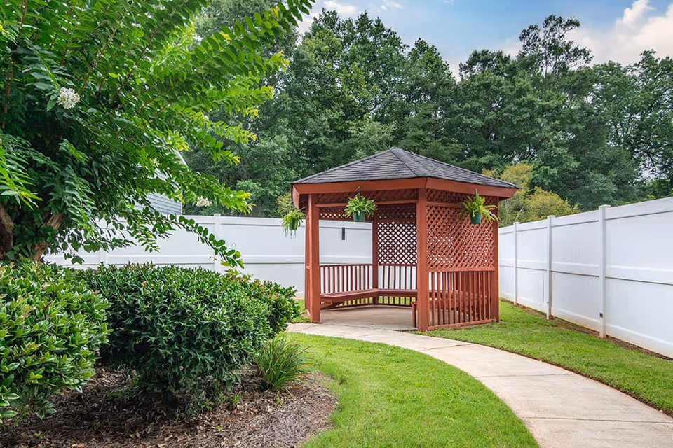 A red wooden gazebo with a shingled roof and hanging green plants is situated at the end of a curved concrete pathway. The gazebo is surrounded by green grass, bushes, and trees, with a white privacy fence enclosing the area.