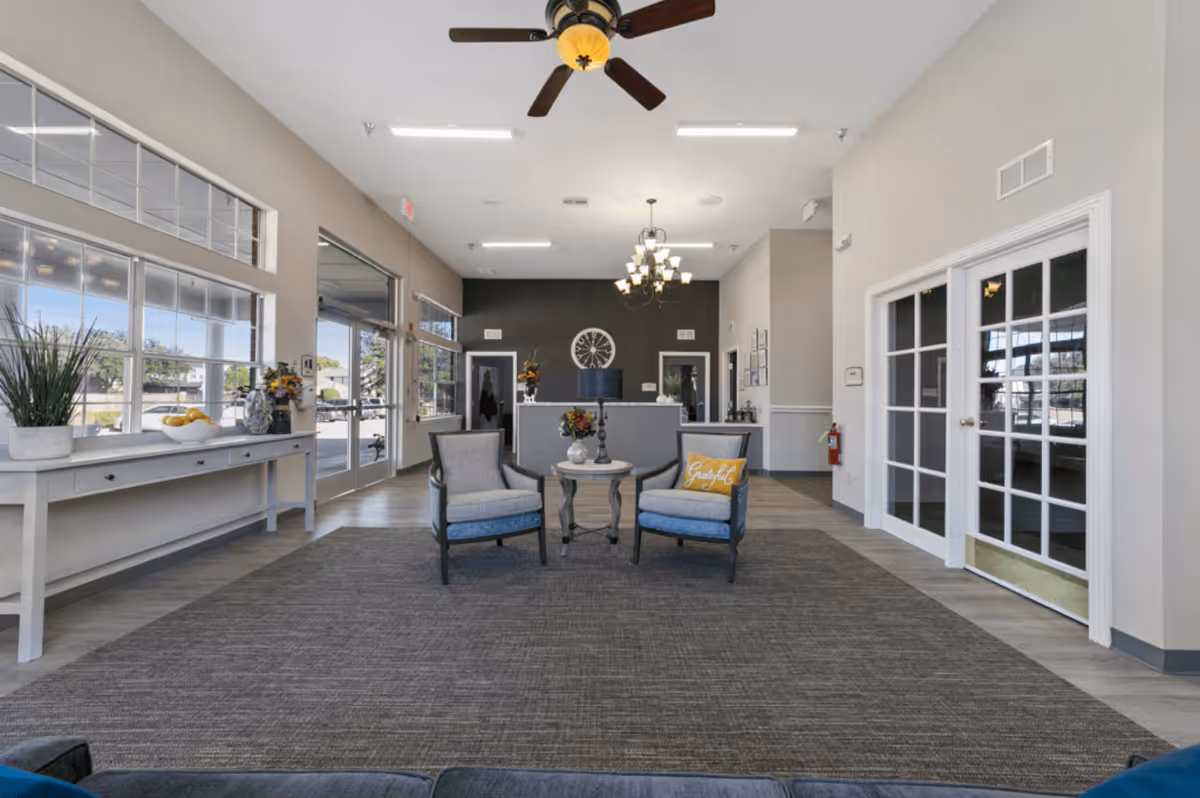 A spacious and well-lit common area in a senior living facility with two armchairs and a small round table in the center on a large gray rug. The room features large windows on the left side, a white console table with decorative plants and a bowl of lemons, and a reception desk against a dark accent wall with a large clock. There are double glass doors on the right and a ceiling fan and chandelier overhead.