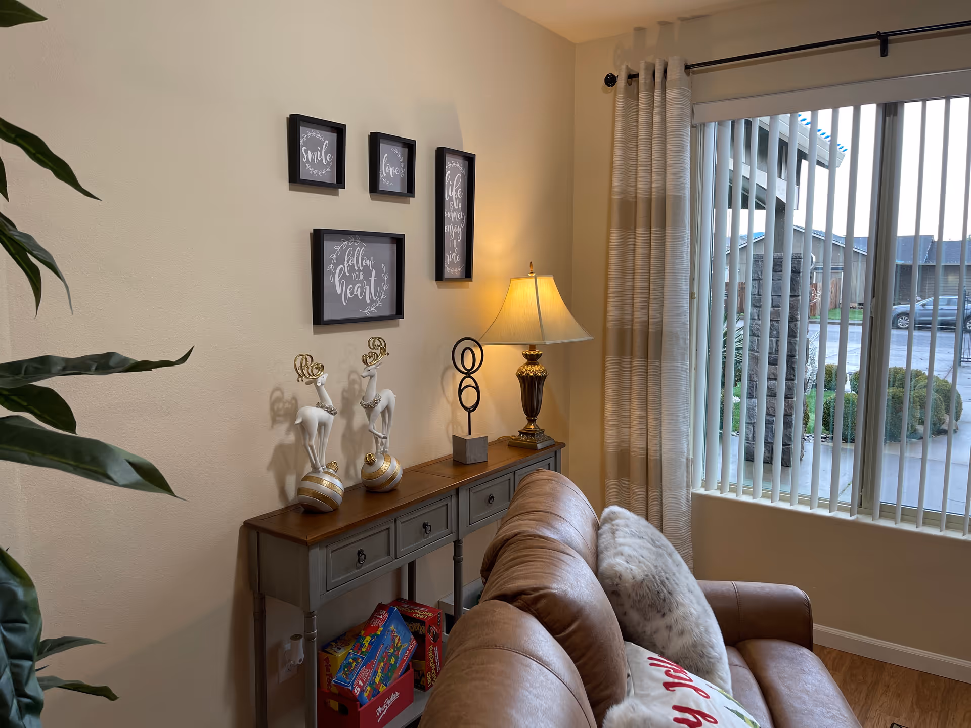 Cozy living room corner with a leather sofa, a console table holding a lamp and decorative figurines, framed wall art, and a window with vertical blinds.