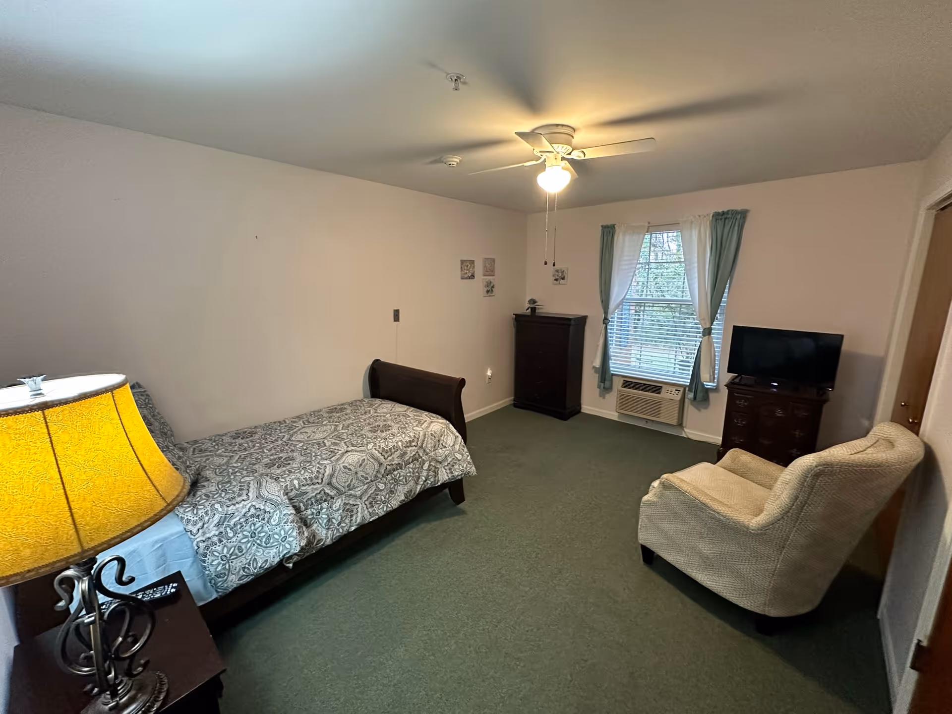 A small bedroom with a single bed, nightstand and lamp, armchair, dresser with TV, and a window with curtains.