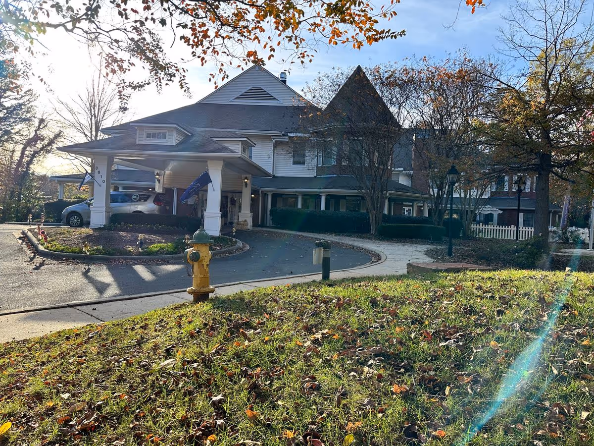 Front entrance and porte-cochère of a senior living facility with a circular driveway, a yellow fire hydrant in the foreground, and autumn trees.