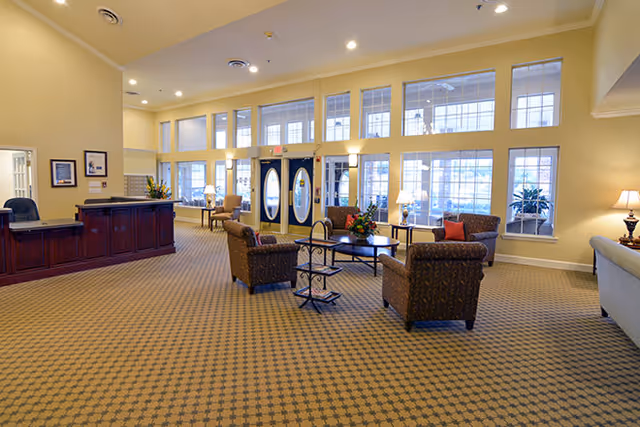 Spacious senior living facility lobby with patterned carpet, a dark wood reception desk on the left, and a seating area with four upholstered armchairs around a coffee table in the center. Large windows and double glass doors allow natural light to fill the room. Lamps and framed pictures decorate the space.