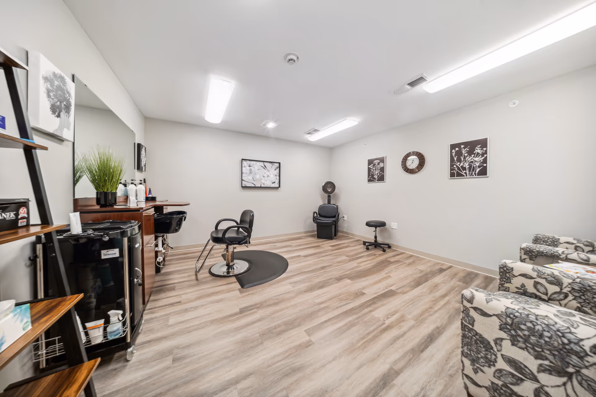 Bright salon/styling room in a senior living community with styling chairs, a shampoo station, shelving, and upholstered seating.