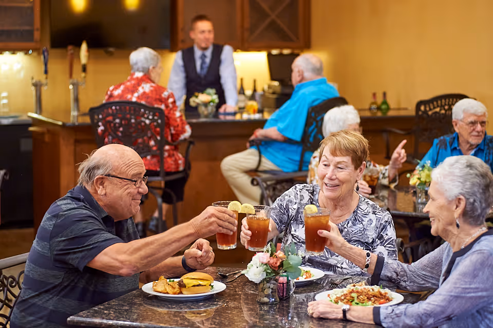 A group of elderly residents clink iced-tea glasses at a dining table in a communal dining area with staff and other residents in the background.