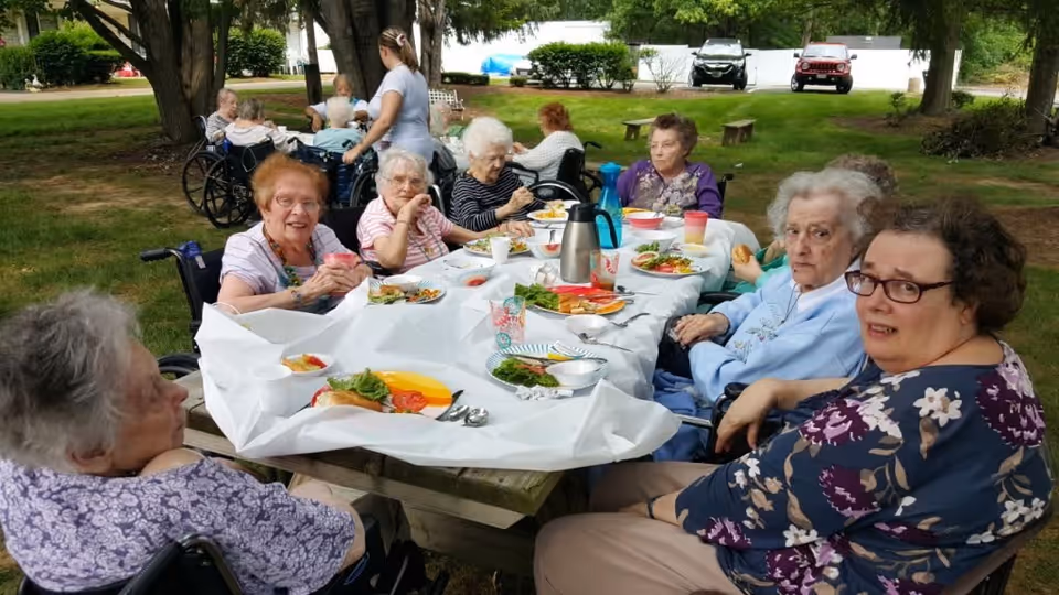 A group of elderly women sitting around a picnic table outdoors, enjoying a meal together. The table is covered with a white tablecloth and has plates of food, cups, and a coffee thermos. Some women are in wheelchairs, and a caregiver is standing nearby. The setting is a grassy area with trees and parked cars in the background.