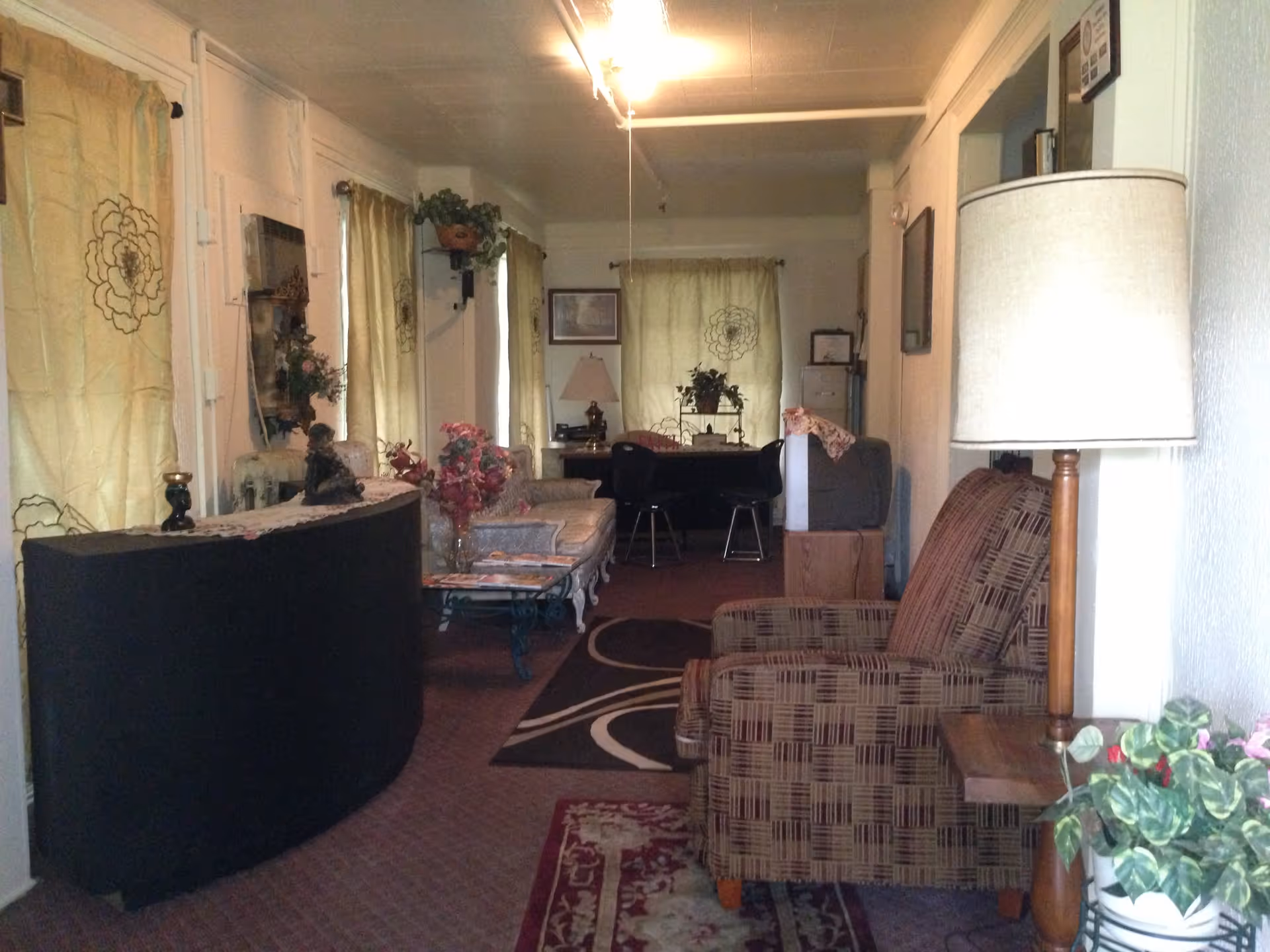 Interior view of a cozy living room area with patterned armchairs, a floor lamp, a small table with a plant, a curved black cabinet, floral curtains, and a sofa with a glass coffee table. The room has carpeted floors and decorative wall hangings.