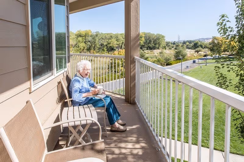 An elderly woman sitting on a chair on a balcony holding a cup, with a small table beside her and a scenic view of green trees and grass in the background under a clear blue sky.
