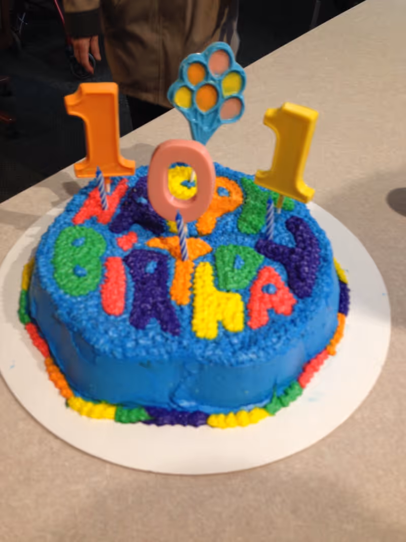 A blue frosted birthday cake decorated with colorful icing letters and candles forming "101" on a table.