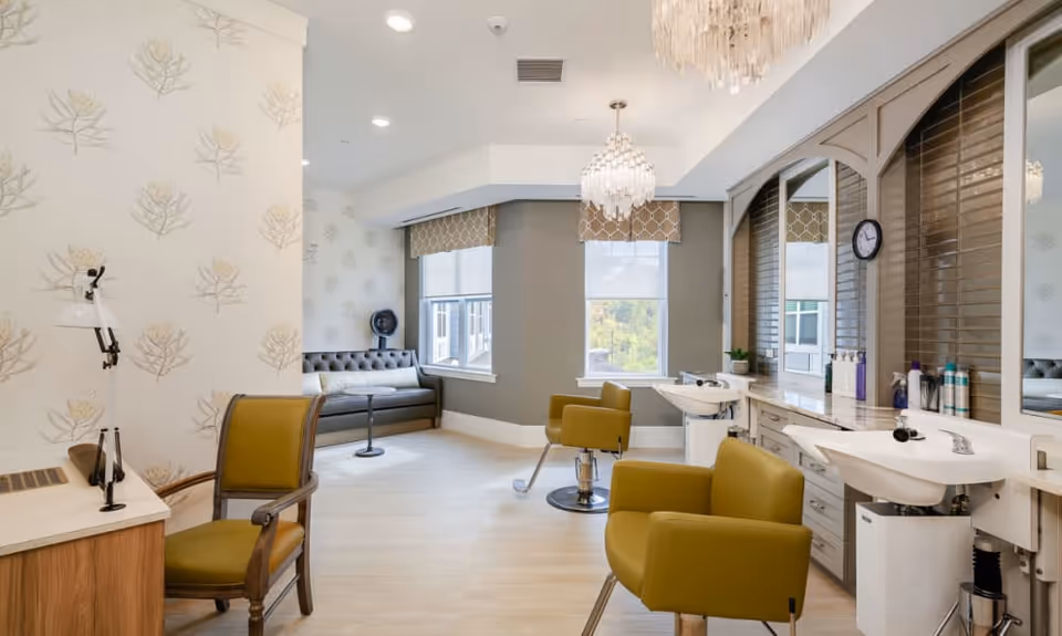 A bright and modern salon area in a senior living facility featuring two olive green salon chairs in front of sinks and mirrors, a tufted gray sofa near windows with patterned valances, a wooden desk with a lamp and chair, and two elegant chandeliers hanging from the ceiling.