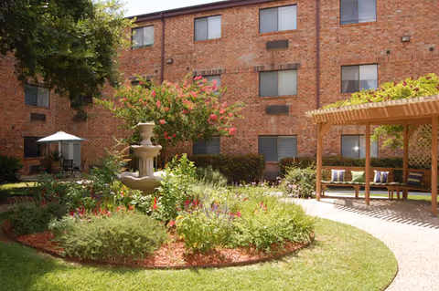 Outdoor courtyard area of a senior living facility with a central fountain surrounded by flowering plants and shrubs. There is a wooden pergola with seating on the right side and a patio umbrella with chairs on the left side. The background shows a three-story brick building with multiple windows.