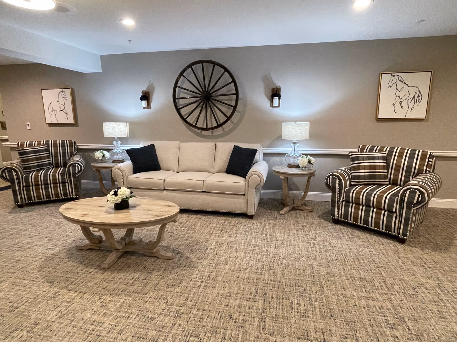 A cozy living room area with a beige sofa adorned with two black pillows, flanked by two striped armchairs. There are two wooden side tables with lamps and small flower arrangements on each side of the sofa. A round wooden coffee table with a small flower arrangement sits in front of the sofa. The wall behind features a large decorative wagon wheel and two framed horse sketches, with two wall-mounted lantern-style lights.