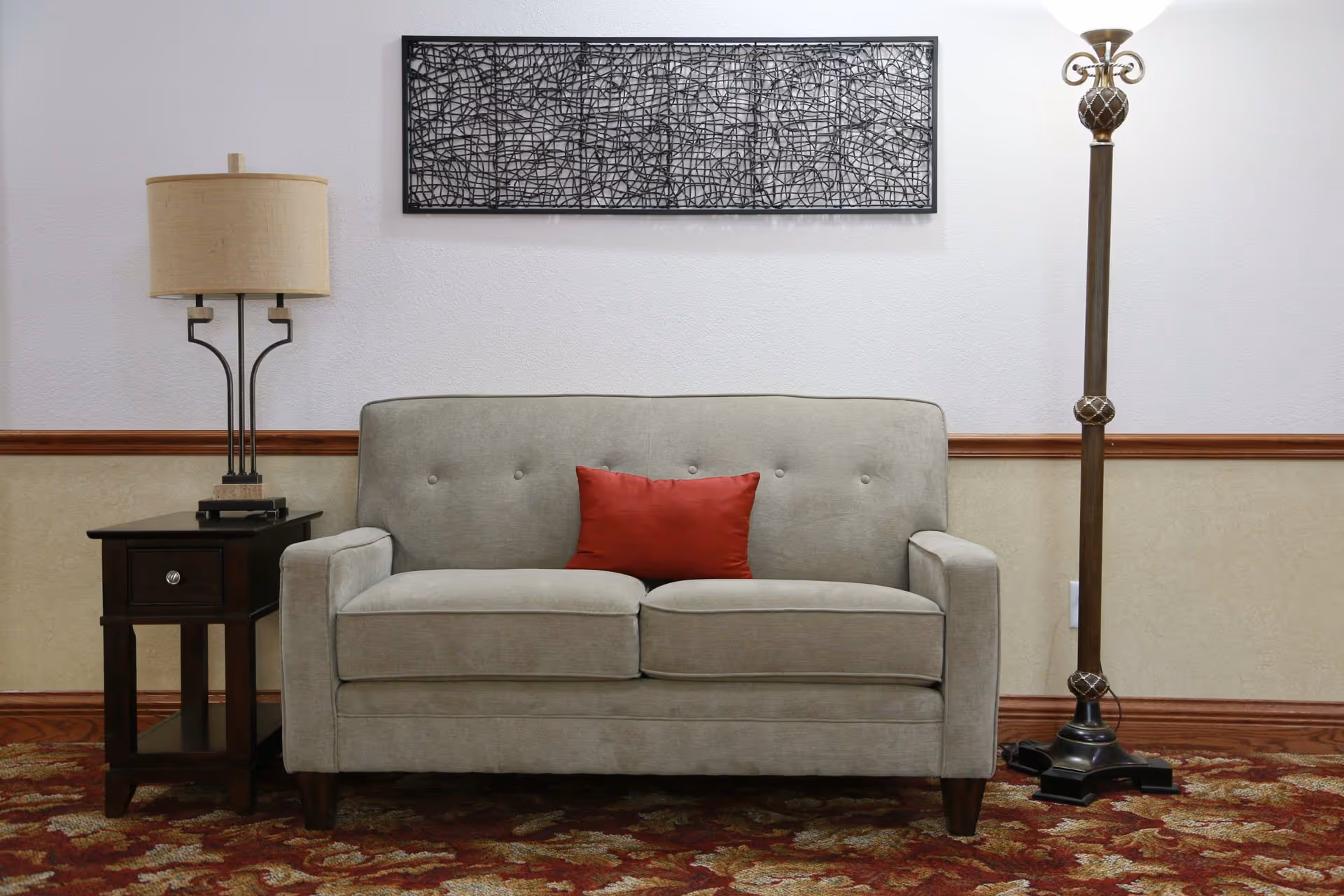 Beige loveseat with a red pillow flanked by a side table and floor lamp beneath wall art in a lounge area.