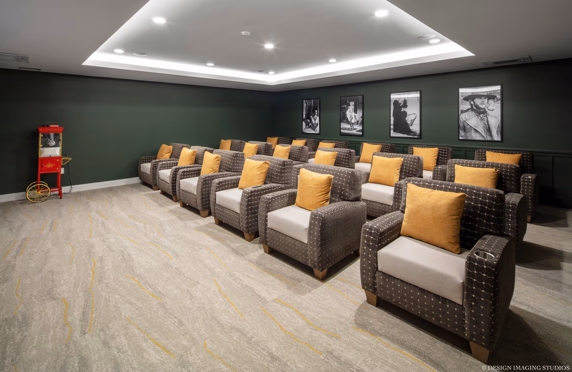 A small theater room with three rows of comfortable armchairs, each with a yellow cushion. The walls are painted dark green and decorated with black and white framed photographs. A red popcorn machine is positioned in the corner of the room.