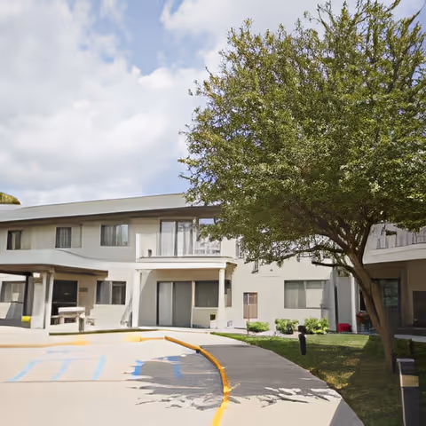 Two-story senior living building with a curved driveway, a tree on a small lawn, and covered ground-floor walkways.